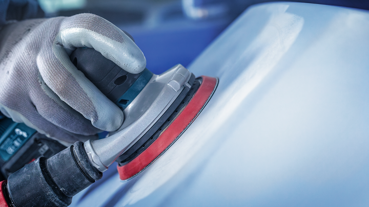 一名佩戴安全设备的人员正在用电动砂光机打磨汽车表面。