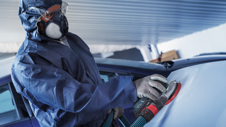 一名佩戴安全设备的人正在用电动砂光机打磨汽车表面。