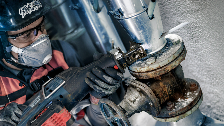 一名佩戴安全设备的人员使用往复锯锯切生锈的管道接头。