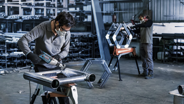 车间里，两个人都在金属框架上使用电动工具，两人都佩戴着安全设备。