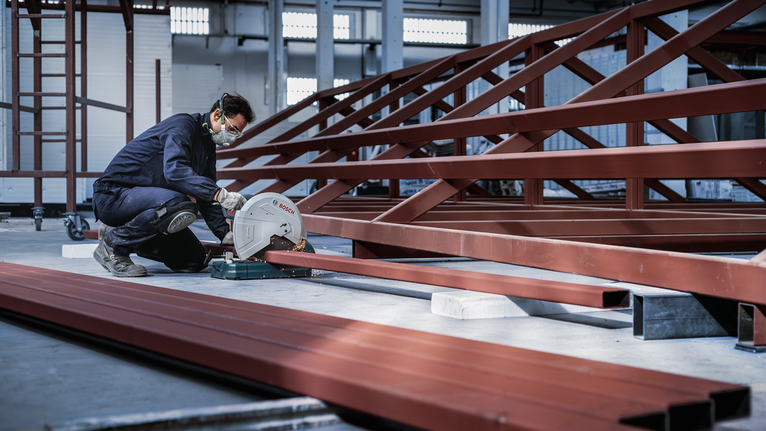 一名佩戴安全设备的工人在工厂里用圆锯切割钢梁。