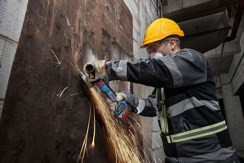一名佩戴安全设备的人正在用无绳角磨机打磨生锈的金属板。.