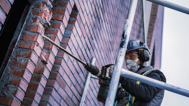 一名身穿安全设备的人员在脚手架上钻入砖墙。
