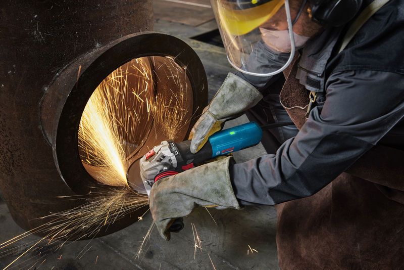 一名佩戴安全设备的人正在研磨一根大金属管，产生明亮的火花。.