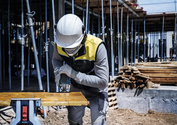 建筑工地上，一名佩戴安全设备的工人正在用电动工具切割木板。