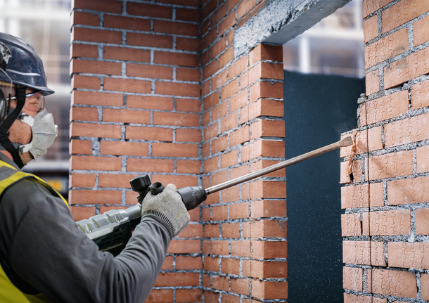 佩戴安全设备的人员使用电动工具凿开砖墙。