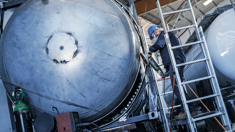 穿着安全装备的人员在梯子上磨大型金属罐的边缘。