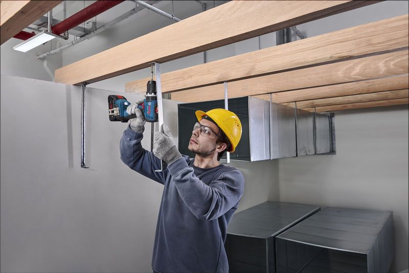 一名佩戴安全设备的人员正在将钻头插入木梁下方的金属棒中。.