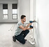 一个人在一间明亮的、未完工的房间里钻孔安装管道。.