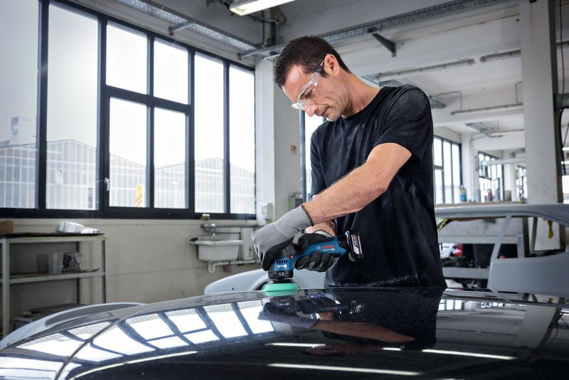 一名佩戴安全设备的人员在汽车修理厂内擦拭汽车引擎盖。.