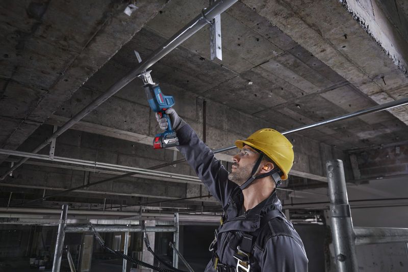 一名佩戴安全设备的人员使用往复锯切割头顶上的金属管道。.