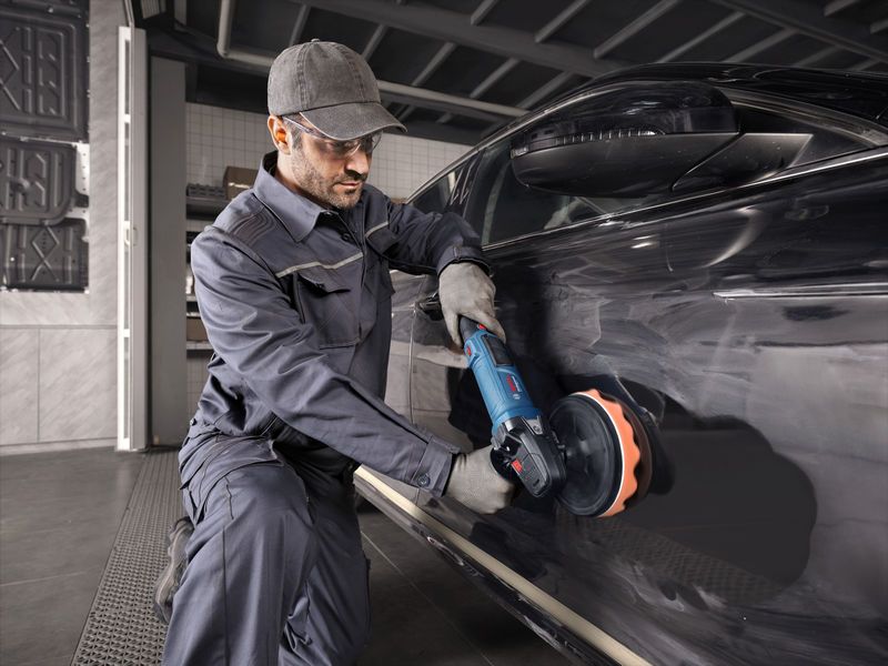 一名佩戴安全设备的人使用电动工具对一辆黑色汽车进行抛光。.