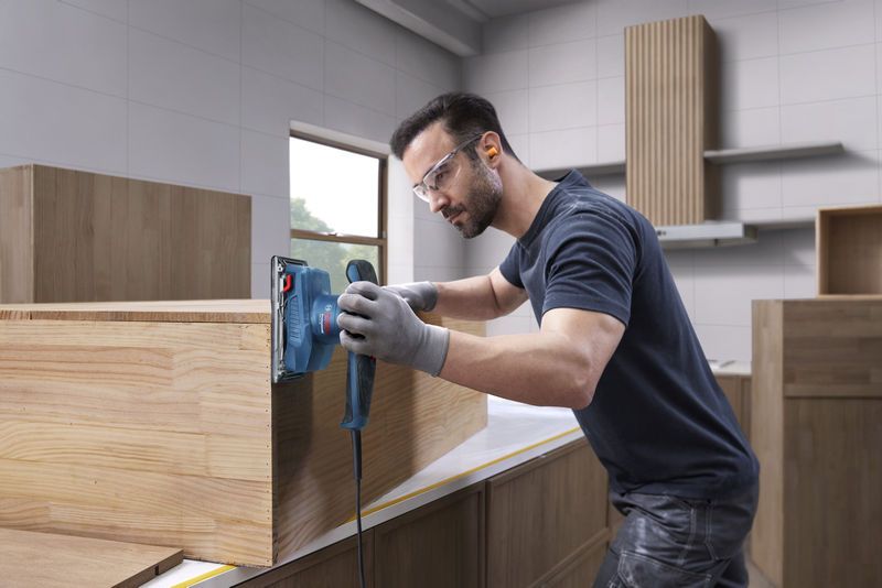 一名佩戴安全设备的人员正在使用轨道砂光机打磨木制橱柜。.