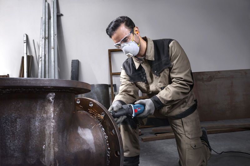 一名佩戴安全设备的人正在磨削一根大金属管的边缘。.