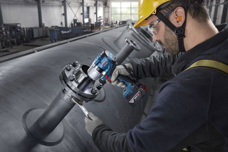 一名佩戴安全设备的人员使用无绳冲击扳手拧紧工业管道上的螺栓。.
