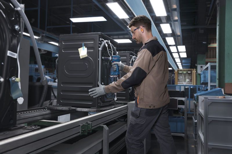 一名佩戴安全设备的人员正在使用电动工具在装配线上固定电器。.