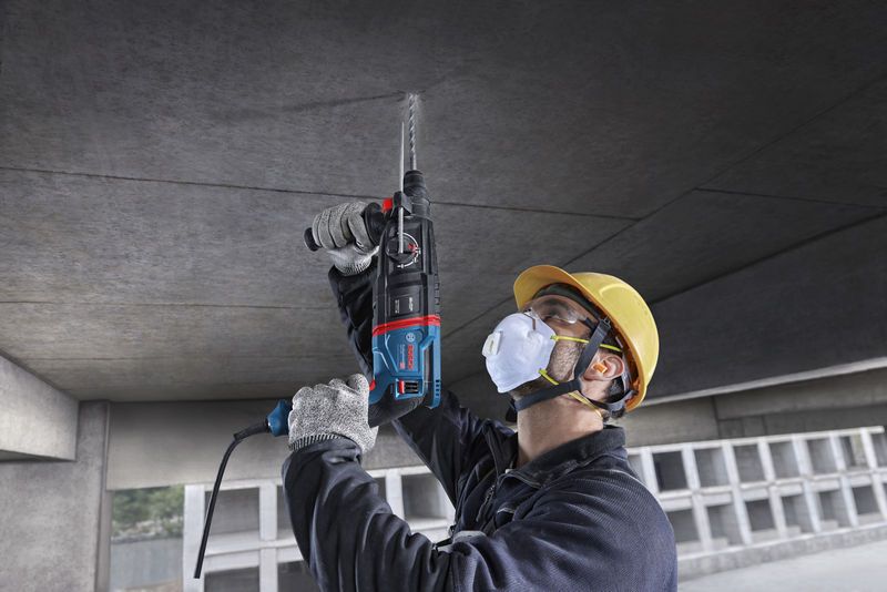 一名佩戴安全设备的人员使用旋转锤在混凝土天花板上钻孔。.