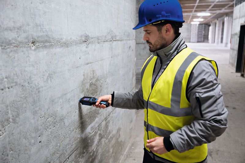 一名佩戴安全设备的人员使用材料湿度计检查混凝土墙体的湿度。.