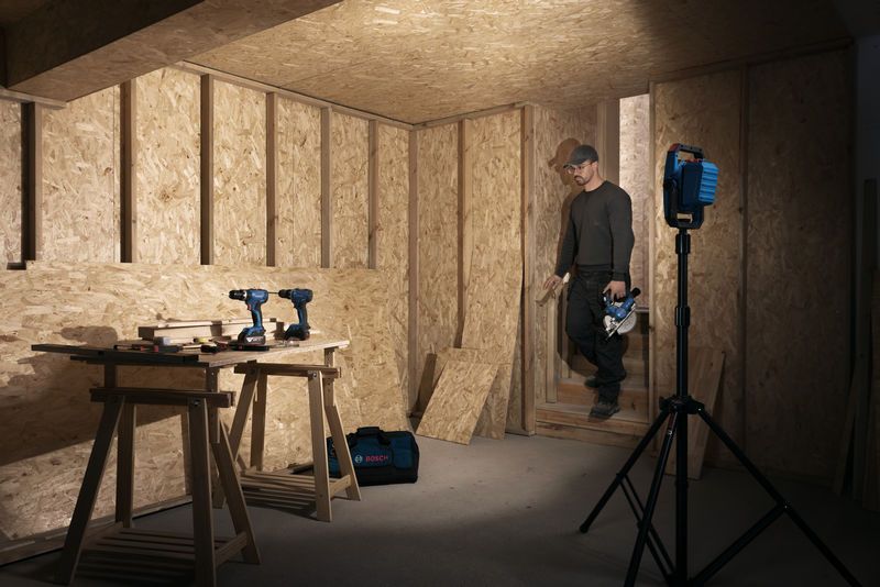 一名佩戴安全设备的人走进由无绳工地灯照亮的木工车间。.