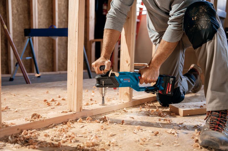 一名佩戴安全设备的人员使用无绳角钻对木框架进行钻孔。.