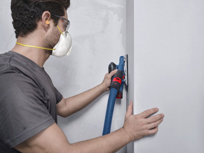 一名佩戴安全设备的人员使用连接到真空软管的轨道打磨机打磨墙壁。.