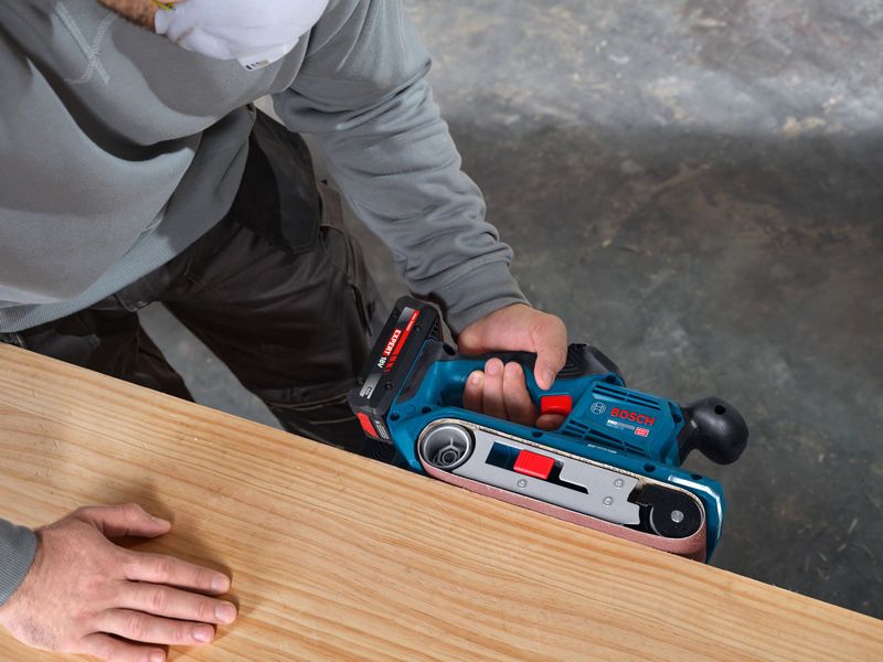 一名佩戴安全装备的人员正在用无绳带式砂光机打磨木板。