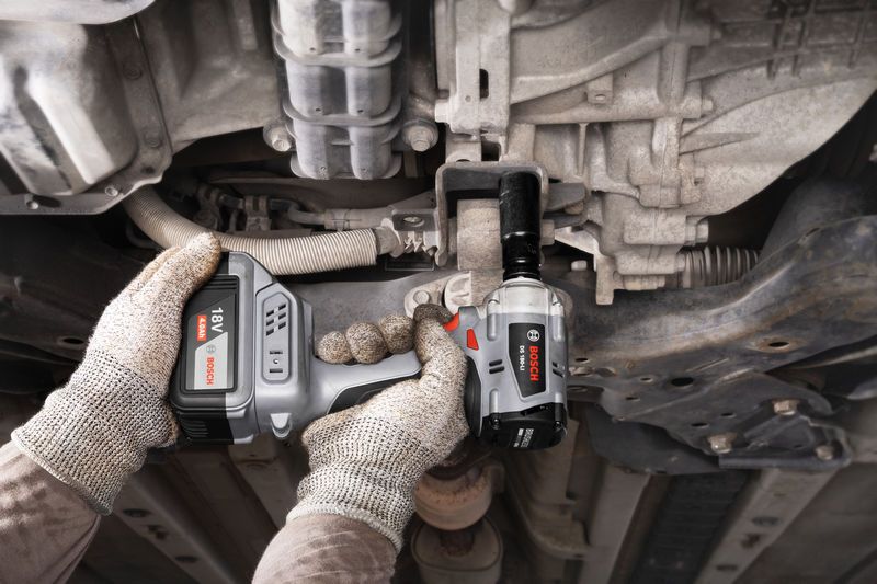 一名佩戴安全设备的人员使用无绳冲击扳手拧紧车辆下方的螺栓。.