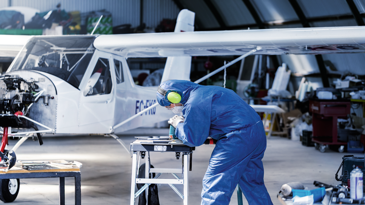 一名佩戴安全设备的人员在小型飞机附近的工作台上钻金属部件。.