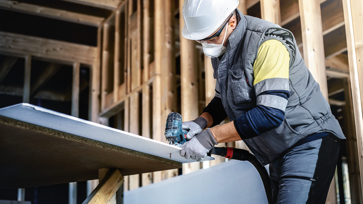 佩戴安全设备的人员使用电钻对建筑框架内的石膏板进行钻孔。.