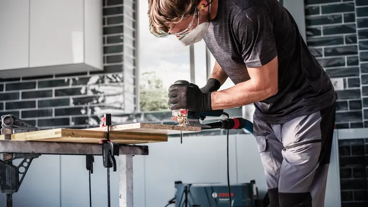 佩戴安全设备的人员使用曲线锯在工作台上切割木材。.