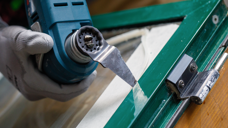 佩戴安全设备的人员使用电动刮刀从窗框上去除密封剂。