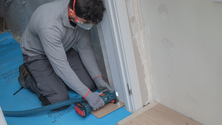 一名佩戴安全设备的人员在门框附近使用电动工具修剪地板。