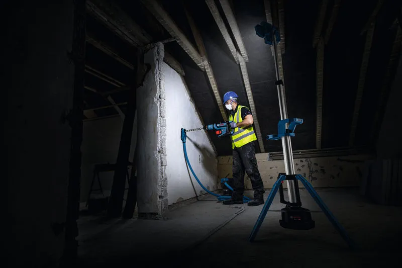 一名佩戴安全装备的人员正在墙上钻孔，一盏无线工地照明灯照亮了墙面。