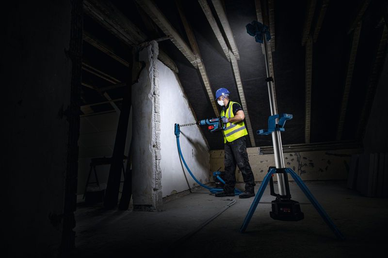 一名佩戴安全装备的人员正在墙上钻孔，一盏无线工地照明灯照亮了墙面。