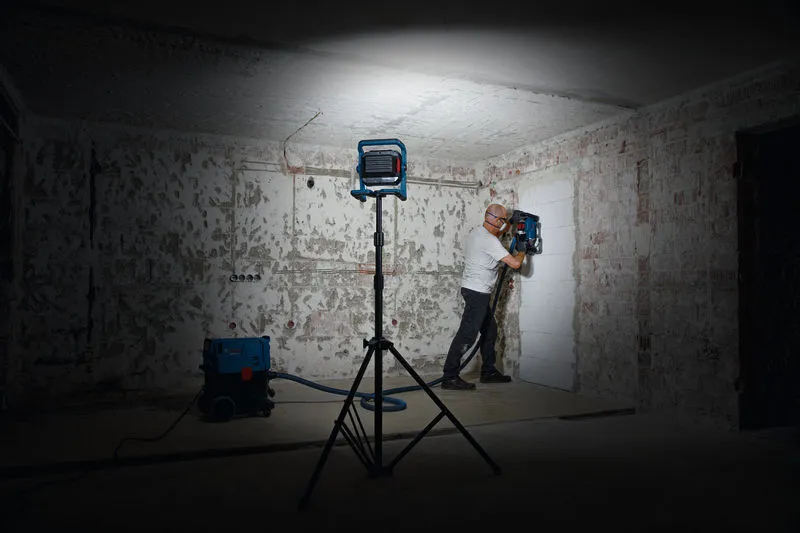 一名佩戴安全设备的人员在工作灯下操作墙壁钻孔工具。