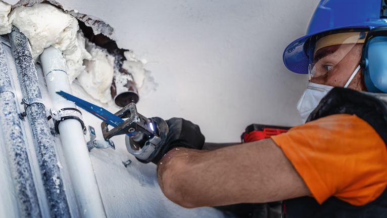 一名佩戴安全装备的人员使用往复锯切割靠近墙壁的管道。