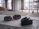 两块可充电工具电池和一个充电器放在建筑工地的水泥地面上。