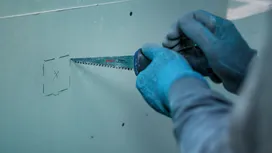 佩戴安全装备的人员用手锯在石膏板上切割出一个标记好的正方形。