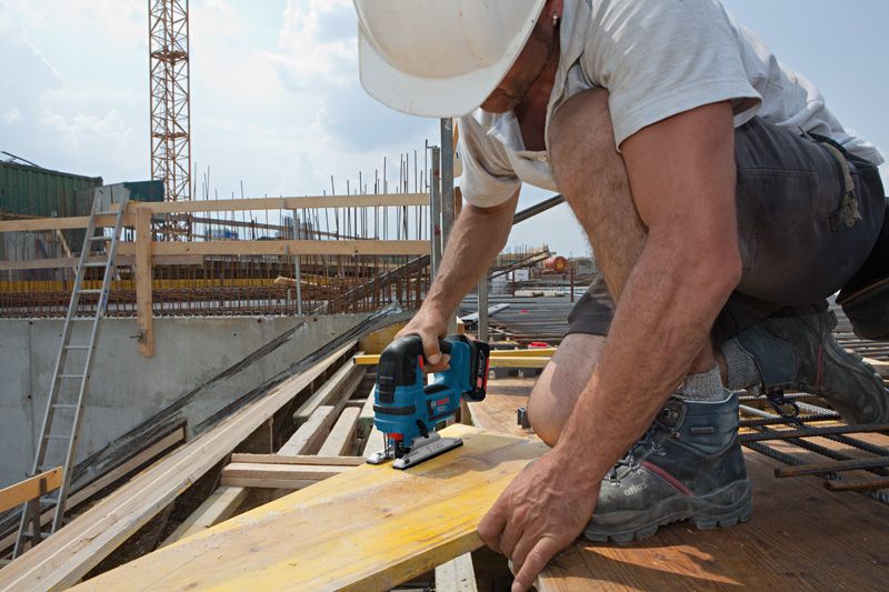 建筑工地上，一名佩戴安全设备的工人正在用无绳曲线锯切割木板。.