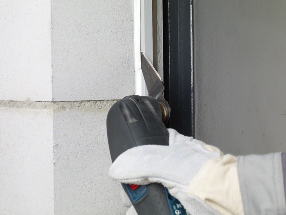 一名穿着安全装备的工人用电动工具从窗框上切下密封剂。.