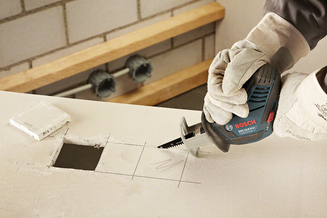 戴着安全设备的工人使用往复锯在干墙上切出一个方形孔。.